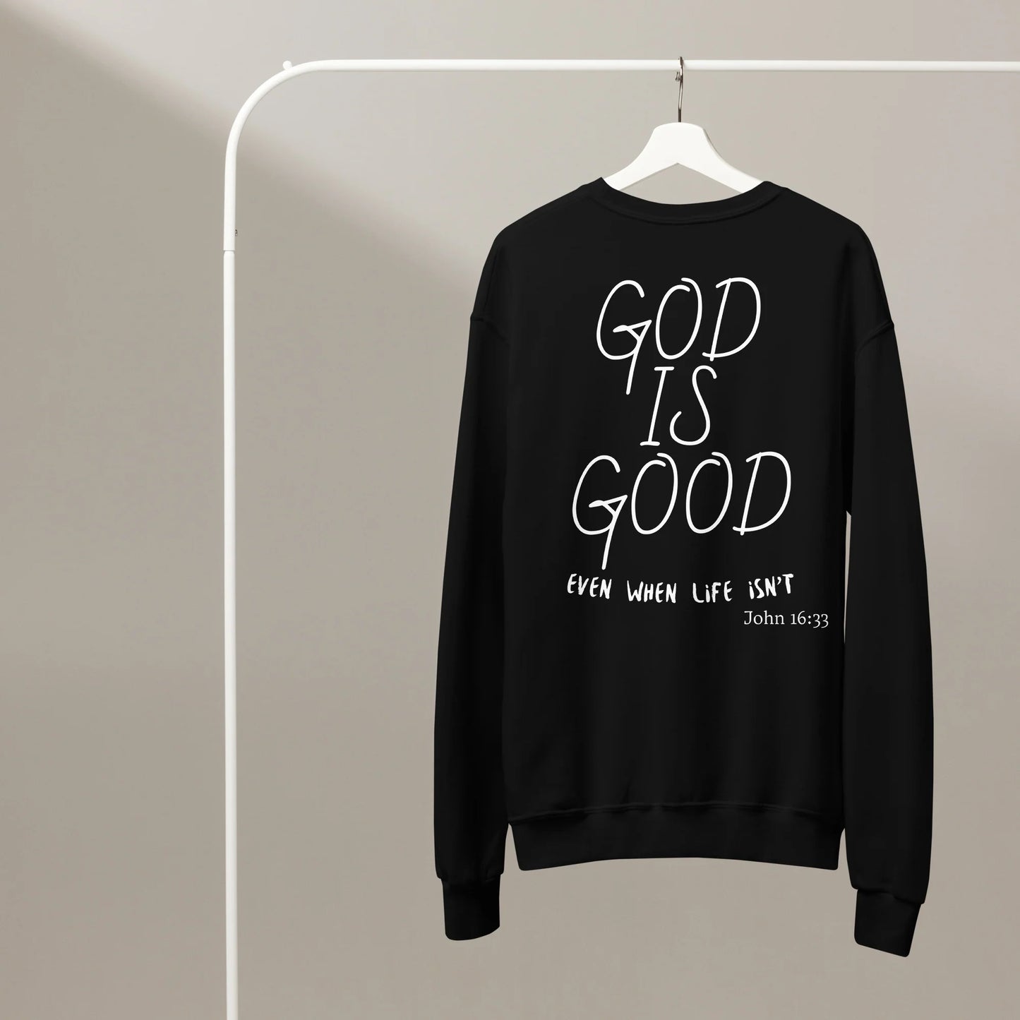 Black sweatshirt with 'God is Good even when life isn't' text on a hanger against a beige background